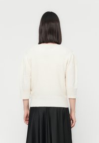 Pull en tricot de couleur crème avec des manches courtes bouffantes et un large décolleté, associé à une jupe noire fluide, vu de dos.