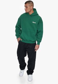 Grøn hættejakke med en frontlomme og hvidt logo, parret med sorte cargo-bukser og hvide sneakers, med en afslappet pasform.
