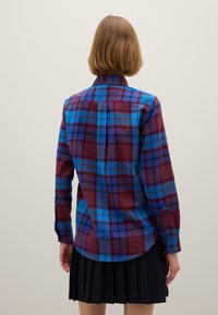 Geruit shirt met blauwe, rode en paarse tinten. Katoenen materiaal, lange mouwen, knoopsluiting. Afgeronde zoom met een ruglijstdetail.