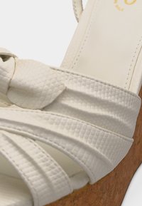 Sandale compensée blanche texturée avec des brides croisées et une semelle en liège, gros plan montrant les détails de la couture et du matériau.