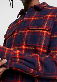 Camicia di flanella blu con motivo a quadri rossi e arancioni, dotata di due tasche frontali, chiusura con bottoni e texture morbida.