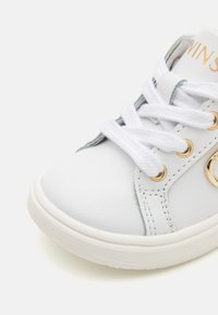 Sneaker sintetico bianco con accenti in metallo dorato, punta rotonda, lacci bianchi e cuciture grigie chiare ai lati. Suola in gomma testurizzata.