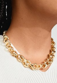 Collana in oro caratterizzata da anelli ovali intrecciati con texture lisce. Indossata su un top di colore chiaro, evidenziando la sua lucentezza.