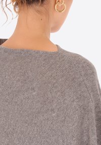 Maglione grigio lavorato a maglia con una vestibilità ampia e rilassata e una texture morbida, caratterizzato da un ampio scollo e dettagli delle cuciture sottili. Orecchini a cerchio dorati visibili.