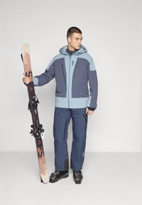 Blue and navy ski jacket with zippers, paired with matching pants. Model holds a ski. Ski boots and ski have a black and red design.