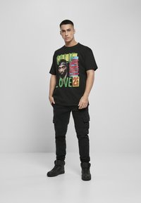 Camiseta gráfica negra con texto colorido y un retrato. Combinada con pantalones cargo negros y zapatillas altas, sobre un fondo simple.