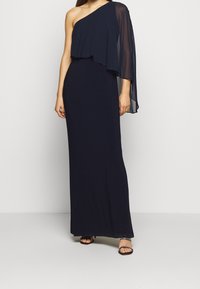 Enaxladad marinblå klänning med flödande chiffongöverdrag, figursydd silhuett och ankellång design. Ett par remsandaler i svart.
