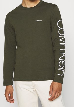 Homme portant une chemise Calvin Klein olive à manches longues avec un logo sur la poitrine et un grand lettrage du logo le long de la manche gauche, associée à un pantalon clair.