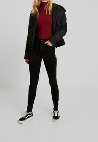 Black quilted jacket, burgundy turtleneck, slim black jeans, and black sneakers with white accents. Neutral background.