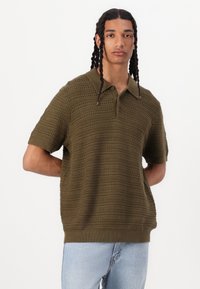 Polo shirt olive vert à manches courtes en tissu tricoté texturé avec un col et un ourlet côtelé, présentant des détails de motif horizontal.