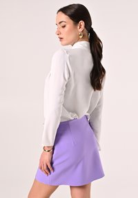 Weiße Langarmbluse, kombiniert mit einem lila Miniröckchen. Die Haare sind offen, und goldfarbene Creolen sind sichtbar. Glattes Material mit einem eleganten Design.