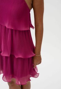 Sukienka w kolorze śliwkowym z plisowanym wzorem, posiadająca warstwowy design i falbaniasty dół. Akcent złotej bransoletki na ramieniu.