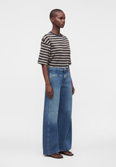 Personne debout de profil, portant une chemise à manches courtes rayée, un jean bleu large et des sandales sur un fond blanc uni.