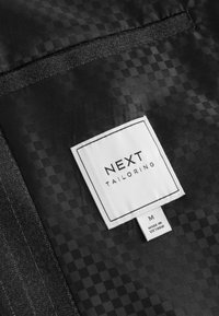 Czarna metka z logo "NEXT TAILORING", z teksturowanym wzorem oraz etykietą z instrukcją pielęgnacji. Rozmiar metki oznaczony jako Medium, wyprodukowano w Wietnamie.