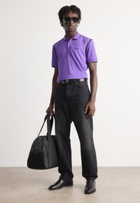 Μωβ πόλο μπλούζα με μαύρες λεπτομέρειες, μαύρα τζην και μαύρα παπούτσια. Κρατάει μια μαύρη τσάντα μεταφοράς. Καθαρό φόντο, στέκεται σε όρθια πόζα.