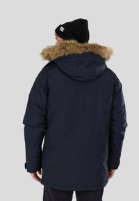 Doudoune marine avec capuche bordée de fausse fourrure, présentant une texture lisse et des poignets élastiques. Portée avec un bonnet noir.