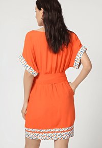 Mujer con cabello oscuro lleva un vestido corto naranja con un ribete blanco decorado en las mangas y el dobladillo, de pie con una mano en el bolsillo.