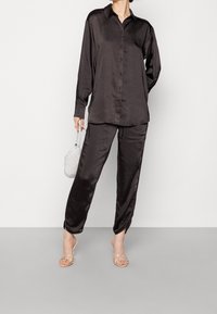 Chemise en satin noir avec fermeture à boutons, assortie à un pantalon. Le mannequin tient un petit sac à main argenté scintillant et porte des sandales à lanières.