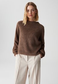 Brauner, flauschiger Pullover mit rundem Ausschnitt und lockerer Passform, kombiniert mit hellbeigen, plissierten Hosen, die ein weiches, strukturiertes Ensemble schaffen.