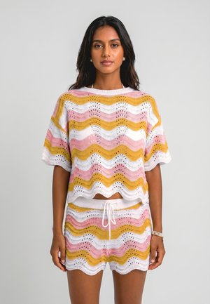 Vrouw draagt een gebreide short en top met golvende strepen in wit, geel en roze en staat tegen een effen lichte achtergrond.