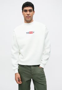 Bílá mikina s kulatým výstřihem, s logem „TOMMY“ v modré a červené barvě. Měkká látka a volný střih, doplněná zelenými kalhotami.