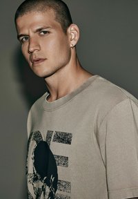 Beżowa, teksturowana koszulka z czarnym nadrukiem przedstawiającym twarz i napisem "ONE". Okrągły dekolt i krótkie rękawy.