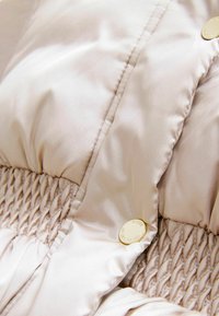 Veste rembourrée beige clair avec une texture lisse et brillante, un design froncé et un accent de bouton doré rond.