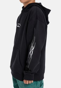 Čierna mikina s kapucňou a prednou vreckom, so striebornými plameňmi na rukáve a logom na hrudi. Mäkká tkanina, voľný strih.