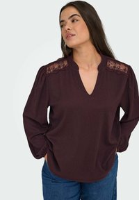 Haut bordeaux avec un décolleté en V, des accents en dentelle sur les épaules, un tissu texturé et de longues manches légèrement ballonnées. Associé à un jean.