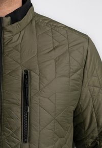 Olivegrüner Steppjacke mit Stehkragen, geprägtem geometrischen Muster und einer Reißverschlusstasche vorne. Leicht und mit glatter Textur.