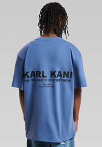 Mėlyna trumpa rankovėmis marškinėliai su ryškiu juodu tekstu: "KARL KANI KATIKROJAS GATVĖS MADOS" ir logotipu. Pagaminta iš medvilnės, laisvo kirpimo.