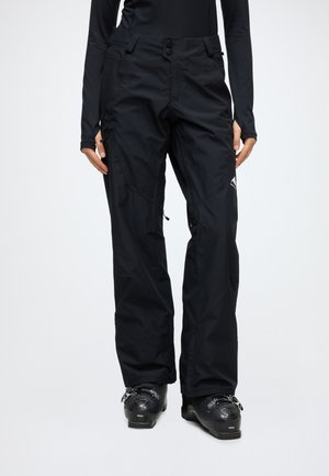 Personne portant un pantalon de ski imperméable noir et des chaussures de ski noires, debout devant un fond clair uni.