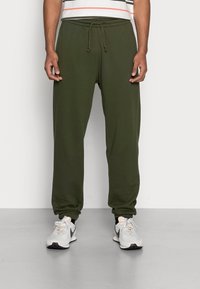 Olijfgroene sweatpants met een trekkoord in de taille, met cuffs bij de enkels, gladde textuur, gecombineerd met grijze sportschoenen op een lichte achtergrond.