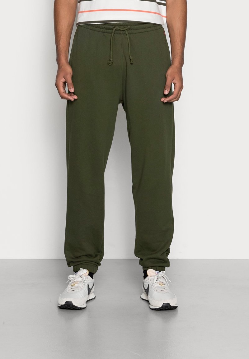 Olijfgroene sweatpants met een trekkoord in de taille, met cuffs bij de enkels, gladde textuur, gecombineerd met grijze sportschoenen op een lichte achtergrond.