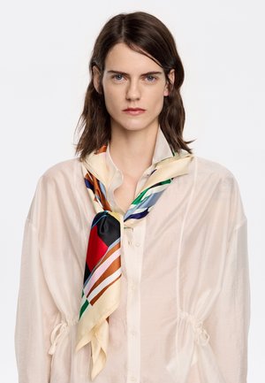 Foulard en soie avec un motif géométrique multicolore, présentant des accents rouges, bleus et crèmes, porté autour du cou sur une blouse beige transparente.