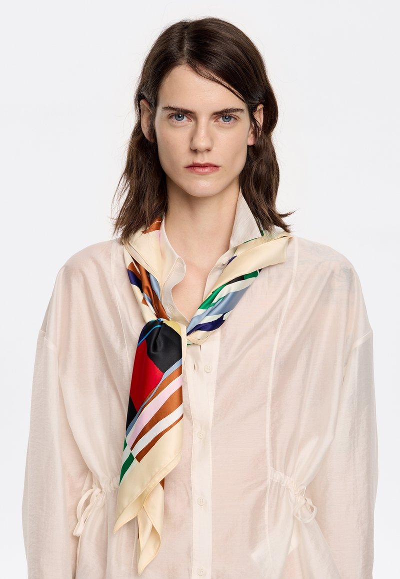 Zijden sjaal met een multicolor geometrisch patroon, met rode, blauwe en crèmekleurige accenten, gedragen om de nek boven een doorschijnende beige blouse.