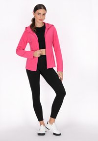 Felpa rosa con zip realizzata in morbido pile, dotata di cappuccio e maniche lunghe. Abbinata a leggings neri e sneakers bianche per un look sportivo.