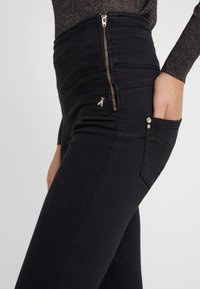 Černé džíny s vysokým pasem, zapínání na zip, ozdobný zlatý motiv včely a přední kapsa se stříbrným detailním knoflíkem. Hladká textura.