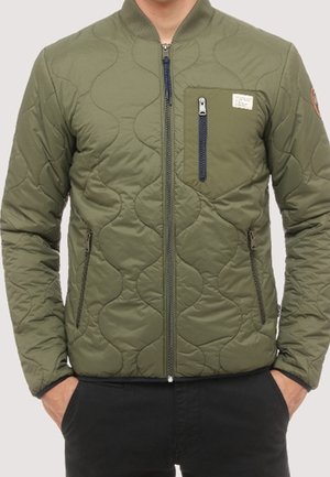 Mann trägt eine grüne gesteppte Jacke mit Reißverschlusstasche auf der Brust und Frontreißverschluss, Hände in den Taschen schwarzer Hosen.