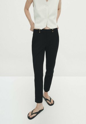 Persoon die zwarte slim-fit broek, wit mouwloos button-up vest en zwarte teenslippers draagt, tegen een effen witte achtergrond.