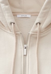 Sweat à capuche beige zippé avec cordons et fermeture éclair argentée, présentant une doublure en polaire douce et une étiquette de marque OPUS sur le col.