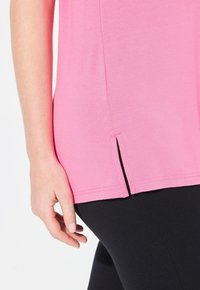 Rosa T-Shirt mit rundem Ausschnitt und Seitenschlitzen am Saum, kombiniert mit schwarzen Leggings. Der Stoff wirkt weich und leicht.