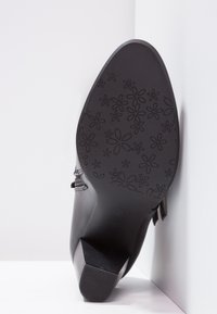 Zapato de tacón negro con una parte superior de cuero suave. La suela presenta un patrón floral, con un diseño de tacón rectangular modesto.