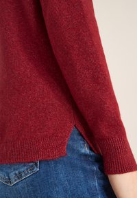 Czerwony sweter z dzianiny, z prążkowanymi mankietami i bocznymi rozcięciami, wykonany z miękkiego materiału. Połączony z niebieskimi dżinsami, widocznymi na dole.