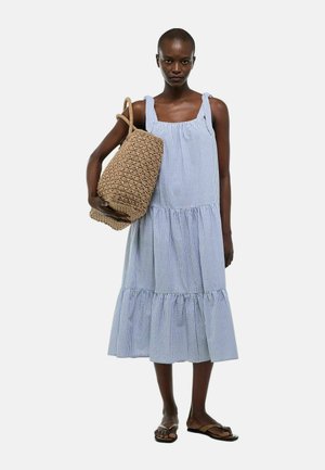 Femme portant une robe midi bleu et blanc à rayures sans manches, sac tressé marron sur l'épaule et sandales marron sur fond uni.