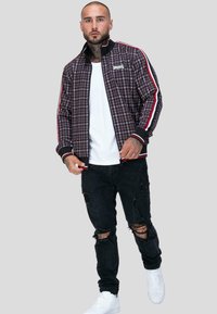 Kurtka bomber w kraciasty wzór w ciemnych kolorach, z czerwonymi i białymi paskami, zestawiona z poszarpanymi czarnymi dżinsami i białymi trampkami.