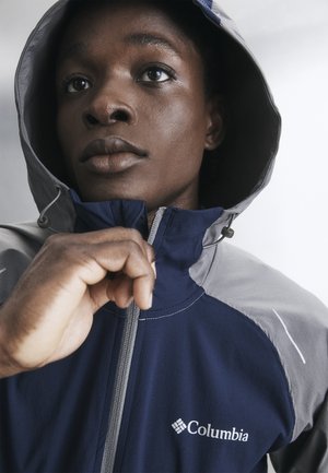 Osoba nosząca granatowo-szarą kurtkę Columbia z podniesionym kapturem, zapinająca ją z przodu, skupione spojrzenie, gładkie jasne tło.