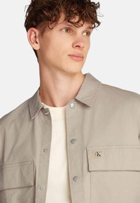 Chaqueta abotonada de color beige claro con una textura suave, dos bolsillos frontales y un pequeño parche con el logo en el pecho. Cuello estándar y cierres a presión.