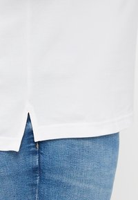 Weißes Hemd mit geradem Saum und seitlichen Schlitzen, hergestellt aus einem glatten Stoff, kombiniert mit blauen Jeans, die eine leicht verblasste Textur aufweisen.