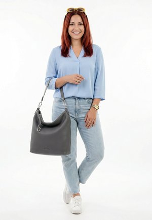 Frau mit rotem Haar, trägt eine blaue Bluse, helle Jeans, weiße Sneaker, hält eine große graue Handtasche, lächelt und hat eine Sonnenbrille auf dem Kopf.